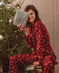 Žena u novogodišnjoj pidžami sedi pored jelke i  drži poklin